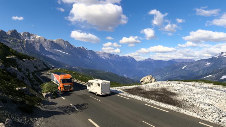 Een oranje truck rijdt op een bochtige bergweg langs een witte camper, onder besneeuwde bergtoppen.